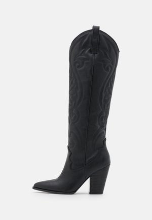 Bottes noires en cuir montant jusqu'au genou avec un embout pointu et un talon bloc. Présente des motifs embossés complexes et une languette en haut.