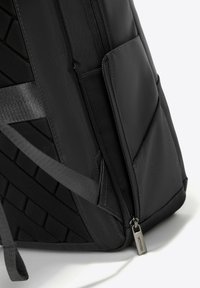 Sac à dos noir en tissu texturé, avec une poche latérale zippée et des bretelles réglables. Le design comprend des motifs géométriques pour un détail supplémentaire.