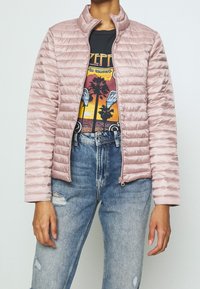 Light pink quilted jacket with a high collar, smooth texture, and zip closure, layered over a graphic T-shirt and distressed blue jeans.