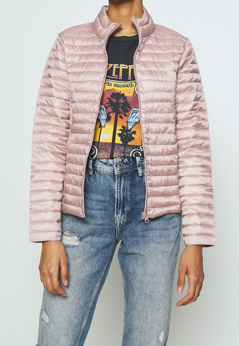 Light pink quilted jacket with a high collar, smooth texture, and zip closure, layered over a graphic T-shirt and distressed blue jeans.
