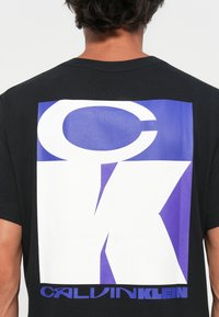 Black t-shirt featuring a large white and purple graphic with "CK" letters and "CALVIN KLEIN" text at the bottom. Standard fit, short sleeves.
