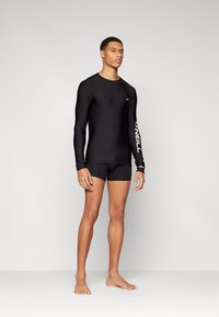 Maglia a maniche lunghe nera con logo sul braccio, abbinata a pantaloni da nuoto neri aderenti. Texture liscia, vestibilità atletica, adatta per sport acquatici.