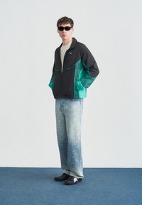 Homme portant des lunettes de soleil noires, une veste Puma noire et verte, un jean bleu clair à motifs, et des chaussures noires, debout sur un sol bleu.