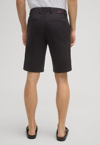 Pantaloni corti neri in cotone con frontale piatto e tasche posteriori, dotati di un'etichetta rossa con logo sulla cintura. L'orlo è rifinito in modo pulito.