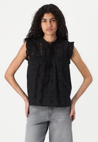 Blusa nera in cotone con maniche a volant, dettagli in ricamo a occhio di pernice e scollatura a chiave, abbinata a jeans grigi.