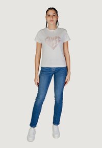 T-shirt bianca in cotone con logo a forma di cuore rosa, abbinata a jeans skinny blu e sneaker bianche. Il modello è in piedi contro uno sfondo chiaro e neutro.