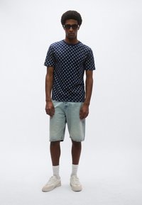 Μπλε μπλούζα με πουά, ανοιχτό μπλε τζιν σορτς, λευκά αθλητικά παπούτσια και κάλτσες κανονικού μήκους. Το μοντέλο στέκεται στο κέντρο μπροστά από ένα απλό φόντο.