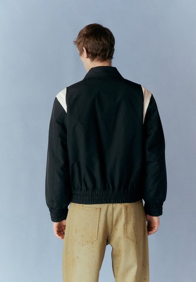 Black jacket with a high collar, elastic waistband, and cream accent on shoulders, paired with light, stained pants. Gray background.