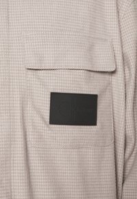 Camicia in tessuto beige chiaro con trama, con taschino applicato e etichetta logo rettangolare nera con la scritta "Calvin Klein Jeans" in rilievo.