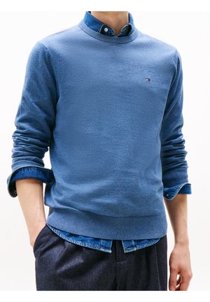 Homme portant un pull bleu clair à col rond sur une chemise en jean retroussée avec un pantalon foncé, mains partiellement dans les poches.