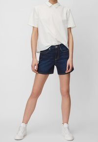 Biała koszula z krótkim rękawem i kołnierzykiem, w połączeniu z ciemnoniebieskimi szortami jeansowymi. Model nosi białe sneakersy za kostkę. Prosty design, brak wzorów.