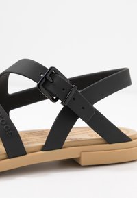 Svart syntetisk sandal med korsettskyddande remmar med matt yta. En svart spänne accentuerar den justerbara remmen. Tan dämpad fotbädd.