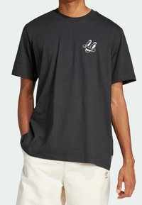 T-shirt en coton noir avec un motif blanc représentant un visage sur le côté gauche de la poitrine. Manches courtes et coupe confortable, associé à un pantalon de couleur claire.