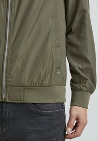 Olivegrüne Bomberjacke mit Reißverschlussfront, gerippten Bündchen und einer Seitentasche. Hergestellt aus leichtem, glattem Stoff mit subtiler Sheen.