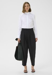 Vit skjorta med puffärmar, kombinerad med högmidjade svarta byxor. Modellen bär en mörkgrön tote-väska och har på sig sandaler.