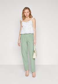 Bílý top s peplum střihem a volánkovými ramínky, s detaily ve tvaru klíčové díry, spárovaný se světle zelenými kalhotami s vysokým pasem a béžovou kabelkou.