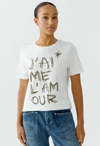 Weißes Baumwoll-T-Shirt mit der Aufschrift "J'ai me l'amour" in goldenen Perlenbuchstaben. Kurze Ärmel, runder Ausschnitt, kombiniert mit blauen Denim-Jeans.