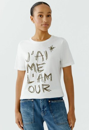 Weißes Baumwoll-T-Shirt mit der Aufschrift "J'ai me l'amour" in goldenen Perlenbuchstaben. Kurze Ärmel, runder Ausschnitt, kombiniert mit blauen Denim-Jeans.
