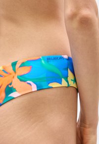 Κοντινό πλάνο στο ισχίο και την κάτω πλάτη ενός ατόμου που φοράει πυτζάμες μπικίνι Billabong με μπλε, πορτοκαλί και πράσινο ανθικό μοτίβο.