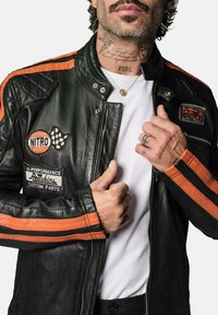 Veste de moto en cuir noir avec des rayures orange, intégrant des écussons et des accents métalliques, fermeture éclair, et un design matelassé sur les épaules.