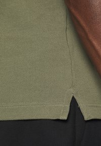 Olivově zelená strukturovaná látka s detailem rozděleného spodního lemu, spojená s černou látkou pod ní. Jednoduchý design s neutrální barevnou paletou.