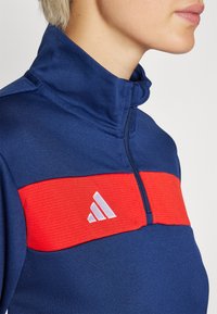 Námornícky a červený pulóver na zips v polovici s hladkým materiálom, vysokým golierom, textúrovanou červenou náprsníkovou časťou a bielym logom na prednej strane.