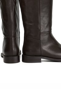 Brown leather knee-high boots with a smooth texture, flat heel, and side zip closure, featuring minimal stitching and a branded logo detail.