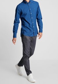 Modrá kostkovaná košile na knoflíky, kombinovaná s tmavě šedými texturovanými kalhotami a bílými teniskami. Košile má límeček a dlouhé rukávy.