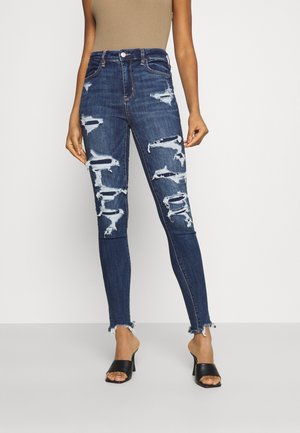 Jeans de tiro alto, de color azul oscuro, desgastados y con detalles rotos en los bordes, combinados con mules negros de punta abierta.