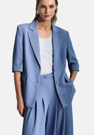 Femme portant un blazer bleu clair avec manches retroussées et un pantalon large assorti, associé à un débardeur blanc côtelé et des boucles d'oreilles dorées.