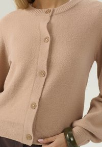 Cardigan beige in lana con bottoni indossato, con bordi a costine e un bracciale verde spesso sul polso sinistro poggiato sui fianchi.