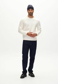 Sudadera de cuello redondo blanca, jeans azul oscuro y zapatos negros. El modelo está de pie contra un fondo blanco, llevando un gorro azul marino.
