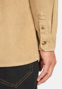 Tan fløjlsskjorte med ribbet tekstur, to-knaps manchetter og lige kant, parret med mørke jeans, der har orange syninger.