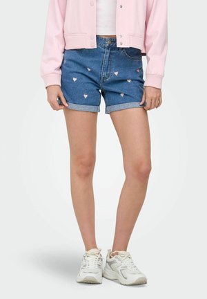 Osoba nosiaca modré rifle krátke nohavice s malými ružovými vzormi srdiečok, biele tenisky, biele tričko a svetloružovú bundu.