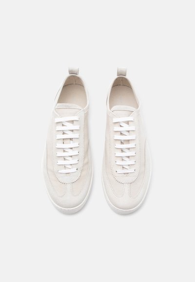 Ζευγάρι εκρού αθλητικά παπούτσια με λευκά κορδόνια, σουέτ διακοσμήσεις και υφασμάτινες θηλιές, σε απλό φωτεινό φόντο.