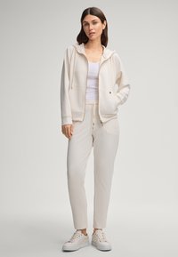 Light beige zip-up hoodie with a hood and front pockets, paired with matching tapered joggers. White t-shirt underneath, minimalistic design.