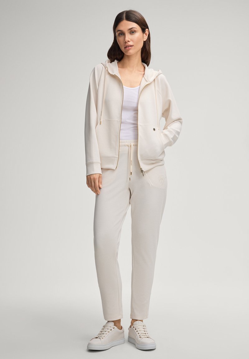 Light beige zip-up hoodie with a hood and front pockets, paired with matching tapered joggers. White t-shirt underneath, minimalistic design.