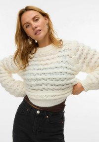 Witte, textuurrijke gebreide sweater met golvende patronen en lange mouwen, gecombineerd met hoog gewaisted zwarte jeans.