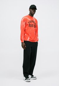 Red tie-dye long-sleeve shirt with black "swamp queen" text, paired with black pants and white sneakers with black accents.