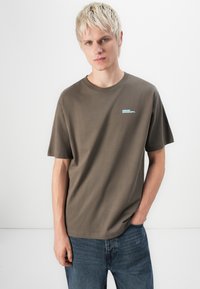 Bruine katoenen t-shirt met een ronde halslijn en korte mouwen, voorzien van een klein lichtblauw logo op de linkerkant van de borst. Draagt in combinatie met blauwe spijkerbroeken.