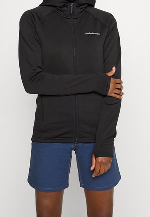 Person wearing a black zip-up hoodie with "Peak Performance" logo and blue shorts, arms crossed, against a plain light background.