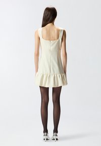 Vestido sin mangas color crema con espalda cuadrada y dobladillo con volantes. Combinado con medias negras y zapatos de tacón alto plateados. Textura suave.