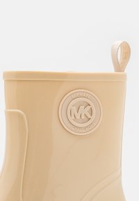Beige rubberen enkellaars met een circulair logo met "Michael Kors." Soepele textuur met een treklus voor gemakkelijk aan- en uittrekken.