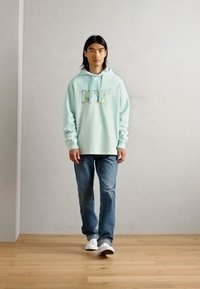 Mintgrøn hættejakke med "Level Up" grafik, parret med blå jeans og hvide sneakers; afslappet pasform, blødt stof og lange ærmer.