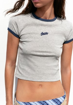 Jeune femme portant un t-shirt court gris clair ajusté avec bordure marine et petit logo marine, associé à un bas de bikini rayé bleu.