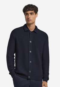 Mörkblå stickad cardigan med ett strukturerat mönster, knäppning framtill och en kontrasterande krage. Har ribbade ärmslut och nederkant.