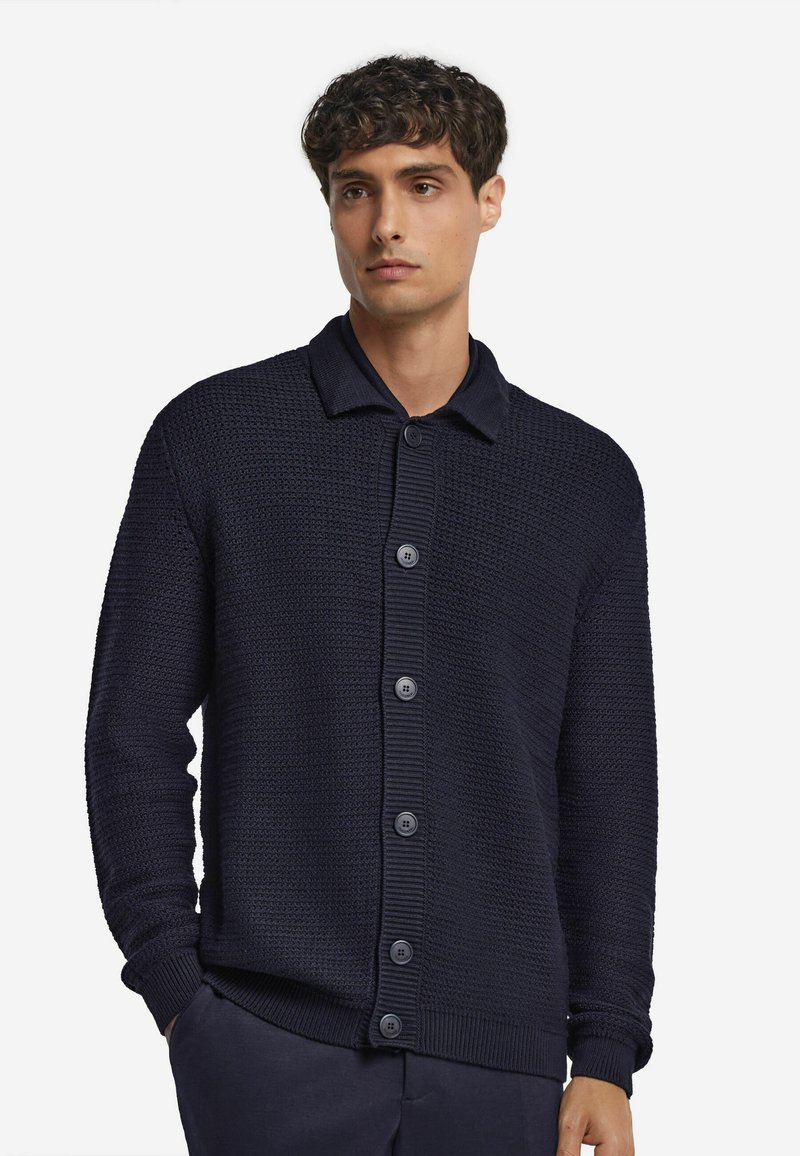 Mörkblå stickad cardigan med ett strukturerat mönster, knäppning framtill och en kontrasterande krage. Har ribbade ärmslut och nederkant.