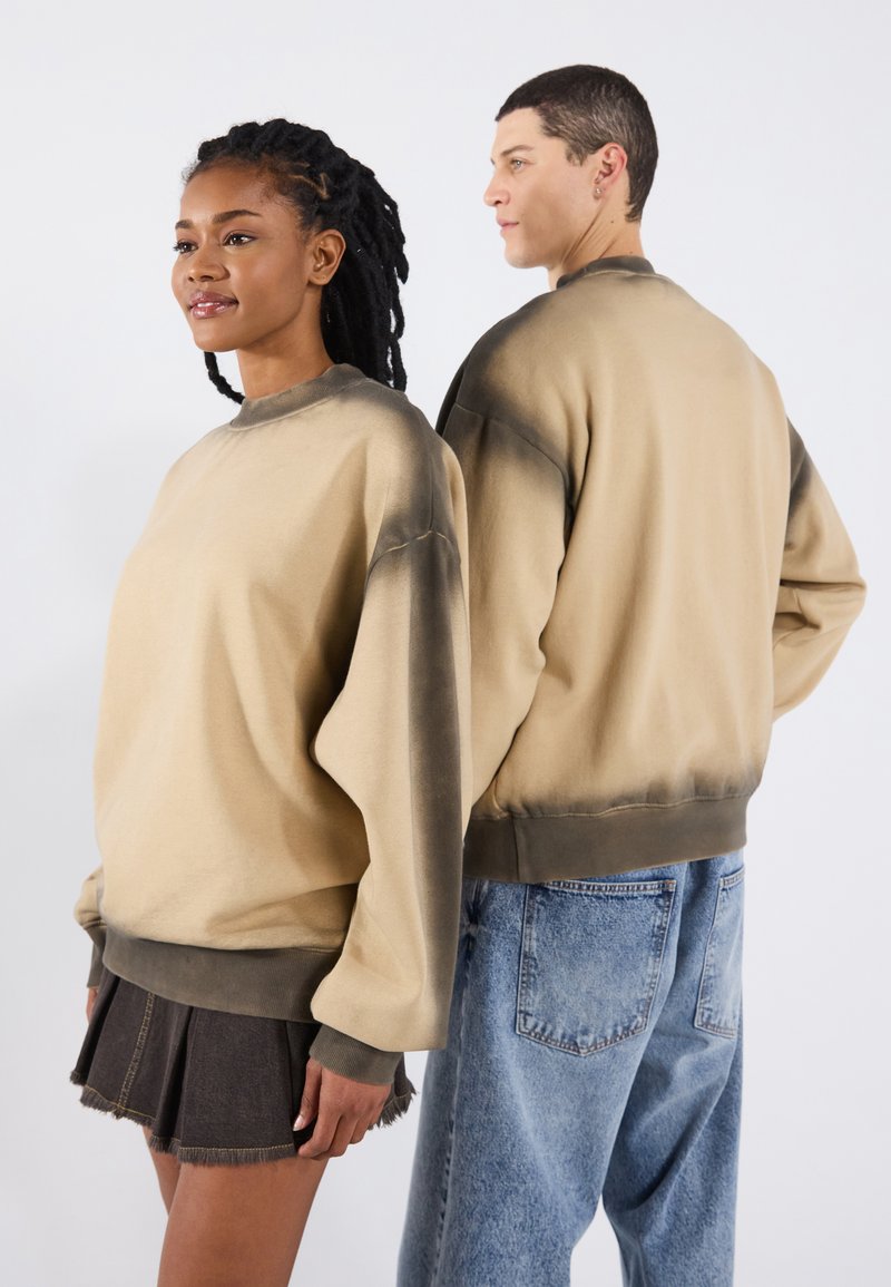 Lichtbruine sweatshirt met een donkere gradient bij de mouwen, ribgebreide halslijn en zoom. Draagt een donkere spijkerrok en lichtblauwe jeans.