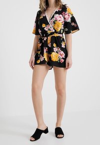 Musta kukallinen rompers, jossa on vaaleanpunaisia ja keltaisia ruusuja, V-pääntie, lyhyet hihat, vyötärölle solmittava vyö ja pitsireunat.