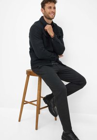 Donkergrijs jack met lange mouwen en knoopklepzakken, gecombineerd met bijpassende broek en zwarte laarzen, zittend op een houten kruk.